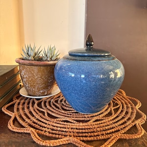May include: A blue ceramic jar with a matching lid featuring a dark pointed finial. The jar has a rounded body and a glossy, speckled finish. It sits on a round, brown, woven fibre mat with a scalloped edge, next to a brown ceramic pot.