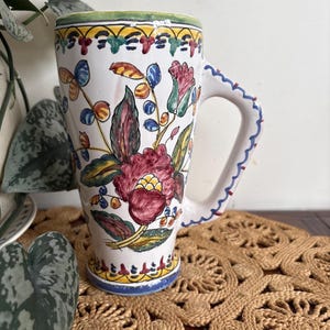 May include: A tall, white ceramic mug featuring hand-painted floral motifs in shades of red, green, yellow, and blue. The design includes a decorative border at the top and bottom, and a sturdy handle with a blue scalloped pattern along the edge.