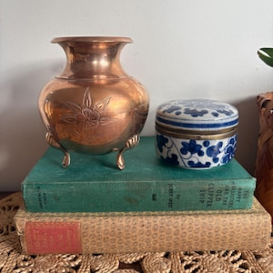 May include: A copper vase with a floral design and three small feet sits on a stack of vintage books. Next to it is a small, round, blue and white ceramic container. The books are green and tan.