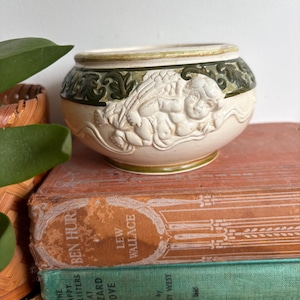 May include: Cream ceramic planter featuring a cherub relief design. The planter has a green and cream rim and base. The cherubs are in a reclining pose. The planter is placed on a stack of vintage books.