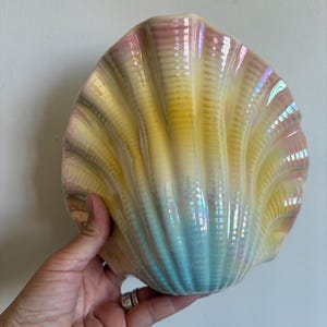 May include: A ceramic shell-shaped bowl featuring a gradient of pastel colours including soft pink, yellow, and light blue. The surface has a glossy, iridescent finish with vertical ridges that mimic the natural texture of a seashell.