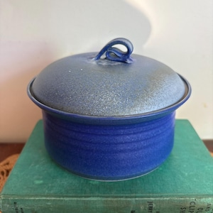 May include: A round ceramic lidded container with a vibrant cobalt blue glaze. The base features subtle horizontal ridges, while the lid has a textured, speckled grey-blue finish and a looped handle at the centre for easy lifting and removal.