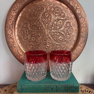 May include: Two glasses with red rims and diamond-cut patterns are displayed on old books. A large, decorative copper tray with floral patterns is in the background. The image evokes a vintage aesthetic.
