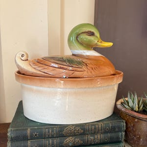 May include: A ceramic casserole dish with a lid shaped like a mallard duck. The lid features a green head, yellow beak, and brown body with detailed feather textures. The base is a round, speckled cream-coloured bowl with a tan rim.