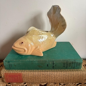 May include: A beige and tan ceramic fish planter with a large tail fin, resting on a stack of vintage books. The books have green and tan covers and are placed on a woven mat. The fish has red eyes.