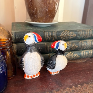 May include: Two ceramic puffin figurines featuring black and white bodies, orange feet, and beaks painted with red, blue, and yellow accents. The taller puffin stands upright while the smaller one is positioned slightly lower with a tilted head.
