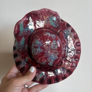 May include: A round, ceramic dish with a ruffled, wavy edge. The surface features a glossy, mottled glaze in shades of deep burgundy, maroon, and patches of light teal blue, creating a variegated, multi-coloured appearance across the entire piece.
