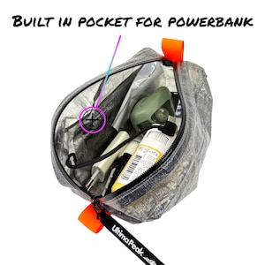 Puede incluir: Una bolsa gris semitransparente con detalles naranjas, llena de varios objetos. El texto "BUILT IN POCKET FOR POWERBANK" es visible en la parte superior. La bolsa tiene una cremallera negra y una etiqueta que dice "UltimaPeak".
