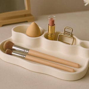 Op de afbeelding: Een crèmekleurige make-up organizer bevat een make-upspons, lippenstift, een gouden haarclip en twee make-up borstels. Een kleine spiegel en een vaas met witte bloemen staan op de achtergrond.