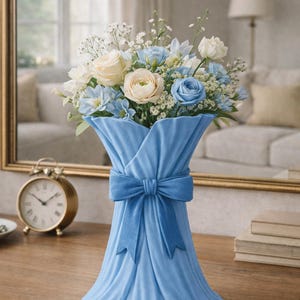 May include: A light blue vase with a bow holds a bouquet of white, cream, and blue flowers. The vase is on a wooden surface, with a gold-colored clock and mirror in the background. The flowers include roses and other blooms.