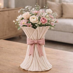 May include: A beige vase with a pink bow holds a bouquet of pink and white flowers. The vase has a draped fabric design. The flowers include roses and other blossoms, creating a soft, romantic arrangement.