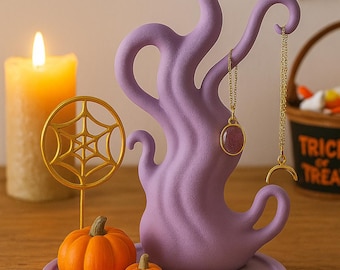 May include: A purple Halloween-themed jewelry holder with a tree-like design, holding necklaces and rings. The display includes orange pumpkins, a gold spider, and a gold web decoration. A lit candle and a Halloween basket are in the background.