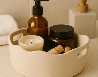 Op de afbeelding: Een beige keramisch dienblad met golvende randen bevat een brandende kaars, een bruine glazen fles met een zwarte pomp en een pot met een zwart deksel. Een kleine houten buis en dop liggen ook in de tray. Een witte handdoek en een dispenser staan op de achtergrond.