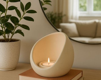 May include: A cream-colored, egg-shaped candle holder with a lit tea light inside, resting on two books. A potted plant and a round mirror are in the background, creating a cozy, minimalist aesthetic.