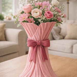 Op de afbeelding: Een roze vaas met een strik bevat een boeket roze en witte bloemen. De vaas heeft een gedrapeerd stoffen ontwerp en een bordeauxrode strik. De bloemen omvatten rozen, madeliefjes en kleine bloesems. De vaas staat op een houten tafel.