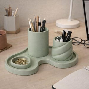 Puede incluir: Un organizador de escritorio verde salvia con tres compartimentos. El organizador contiene bolígrafos, lápices y clips. Una taza de café, un portátil y un cuaderno también están en el escritorio.