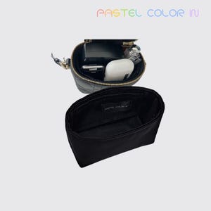 Puede incluir: Un bolso bandolera negro acolchado con cremallera dorada, abierto para revelar su contenido, incluyendo un teléfono, auriculares y otros artículos. Debajo del bolso hay un organizador de tela negra, con el texto "PASTEL COLOR IN".