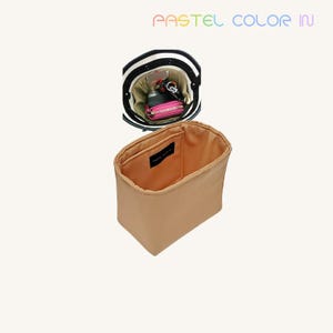K&ouml;nnte beinhalten: Ein beigefarbener Handtaschen-Organizer mit rechteckiger Form und kleiner Tasche. Der Organizer wird neben einer schwarz-wei&szlig; gestreiften Handtasche mit einem rosa Portemonnaie und Schl&uuml;sseln im Inneren gezeigt. Der Text "PASTEL COLOR IN" steht oben.