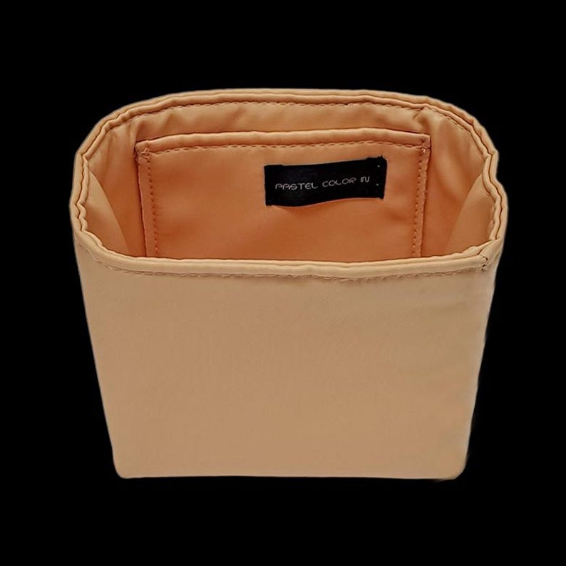 K&ouml;nnte beinhalten: Ein beigefarbener Organizer mit offenem Deckel und rechteckiger Form. Der Innenraum ist in einem helleren Beigeton gehalten und hat eine kleine Tasche. Der Text "PASTEL COLOR N" ist auf einem schwarzen Etikett im Inneren aufgedruckt.