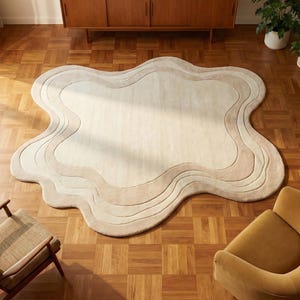 May include: An overhead shot of a cream-colored, wavy-edged rug with a layered design in shades of beige and tan. The rug is placed on a parquet floor, with a mid-century modern style wooden cabinet and two armchairs in the background.