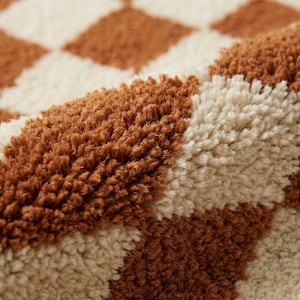 Puede incluir: Primer plano de una alfombra afelpada con un patr&oacute;n repetitivo de rombos marrones y crema. La alfombra tiene fibras gruesas y texturizadas, creando una apariencia suave y acogedora. La paleta de colores es c&aacute;lida y neutra.