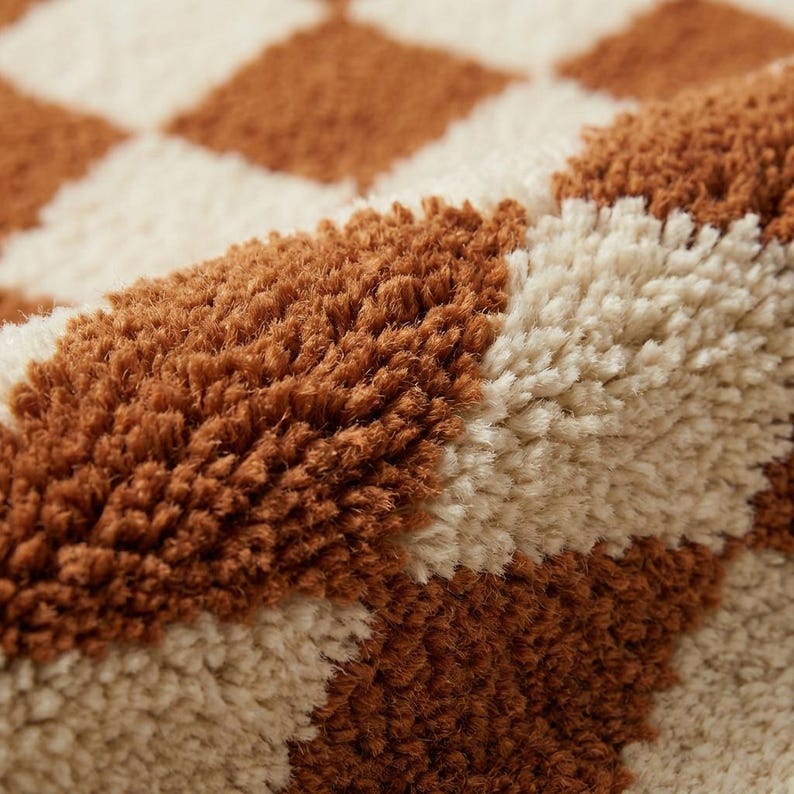 Puede incluir: Primer plano de una alfombra afelpada con un patr&oacute;n repetitivo de rombos marrones y crema. La alfombra tiene fibras gruesas y texturizadas, creando una apariencia suave y acogedora. La paleta de colores es c&aacute;lida y neutra.