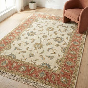 Può includere: Tappeto rettangolare color crema con bordo color ruggine. Il tappeto presenta un motivo floreale nei toni del blu, oro e verde. Il tappeto ha una frangia sui lati corti. Una sedia è sullo sfondo. Ideale per il soggiorno.