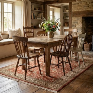 May include: A rustic dining room scene featuring a wooden table surrounded by six wooden chairs. A floral arrangement sits in a vase on the table. A red patterned rug covers the floor, and the room has a warm, inviting atmosphere.