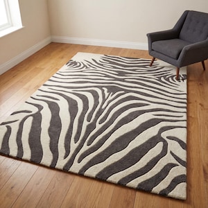 Op de afbeelding: Een rechthoekig vloerkleed met een zebra streepmotief in grijstinten en gebroken wit. Het kleed ligt op een houten vloer, met een grijze fauteuil op de achtergrond. Het ontwerp van het kleed voegt een gedurfd, grafisch element toe aan de kamer.