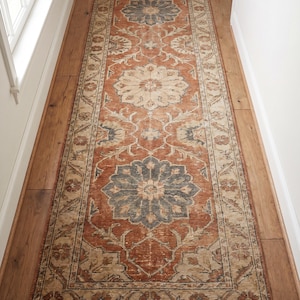 Könnte beinhalten: Ein Läufer im antiken Stil mit einem verblichenen Blumenmuster in Rost-, Beige- und Blautönen. Der rechteckige Teppich liegt auf einem Holzboden und verleiht dem Raum Wärme und Textur. Die Abmessungen des Teppichs betragen ca. 90 cm Breite und 240 cm Länge.