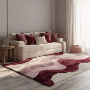 Könnte beinhalten: Wohnzimmer mit cremefarbenem Sofa und weinroten Kissen. Ein großer Teppich mit fließendem, abstraktem Design in Weinrot, Rosa und Creme. Ein kleiner Beistelltisch aus Holz und ein rosa Hocker sind ebenfalls zu sehen.