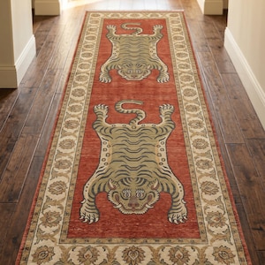 May include: A long rectangular runner rug featuring a vibrant terracotta red background with two stylised, light olive green tiger motifs. The rug is framed by a wide, cream-coloured border decorated with intricate floral patterns and has fringe.