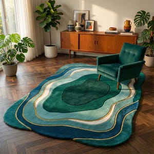 May include: A large, abstract area rug with a layered design in shades of teal, green, and white, accented with gold trim. The rug has an organic, flowing shape and is placed on a hardwood floor. A teal armchair sits on the rug.
