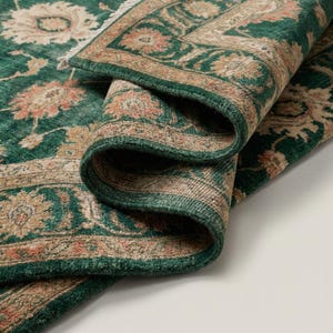 May include: A rolled-up, green area rug with a detailed floral pattern in shades of beige and peach. The rug's border is a darker green, and the edges are slightly frayed, giving it a vintage look. The rug is folded over, revealing its underside.