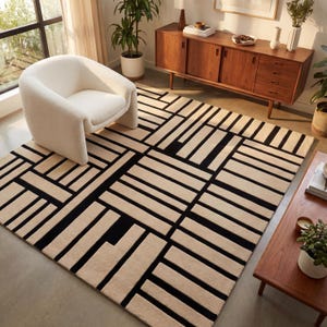 May include: A beige and black geometric patterned area rug. The rug features a repeating pattern of rectangular shapes in beige and black. The rug is placed in a living room setting with a white chair and wooden furniture.