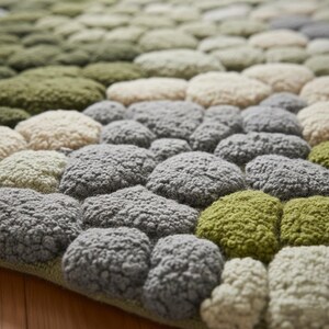May include: A textured rug with a pebble design. The rug features a variety of colors, including shades of green, gray, and cream. The design is made up of rounded, raised shapes, creating a 3D effect. The rug is likely made of a soft material.