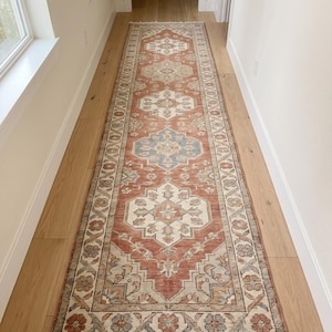 Può includere: Un lungo tappeto passatoia rettangolare con sfondo rosso terracotta e medaglioni geometrici ripetuti in tonalità crema, azzurro e beige. Il tappeto presenta un bordo decorativo con motivi floreali e frange bianche su entrambe le estremità.