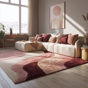 May include: A modern living room featuring a cream-colored sectional sofa adorned with various shades of red and pink throw pillows. A large area rug with a wavy pattern in similar colors complements the sofa. A framed abstract artwork hangs on the wall.