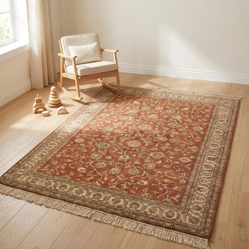 May include: A rectangular, rust-colored rug with an intricate floral pattern and a cream-colored border. The rug has a fringed edge and is placed on a light-colored wooden floor. A wooden rocking chair and toys are in the background.