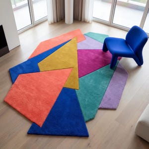 May include: A colorful geometric rug with a modern design. The rug features various shapes in shades of orange, blue, yellow, pink, teal, and purple. A blue chair sits nearby. The rug is on a light wood floor.