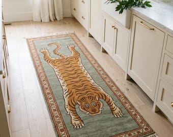 May include: A rectangular runner rug featuring a sage green background with a central illustration of a tiger in shades of orange, black, and white. The rug has a decorative border with floral motifs and white fringe on both short ends.