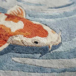 May include: Close-up of a textured rug featuring a koi fish design. The fish has orange and white markings, set against a backdrop of blue and gray water patterns. The rug's texture is visible.