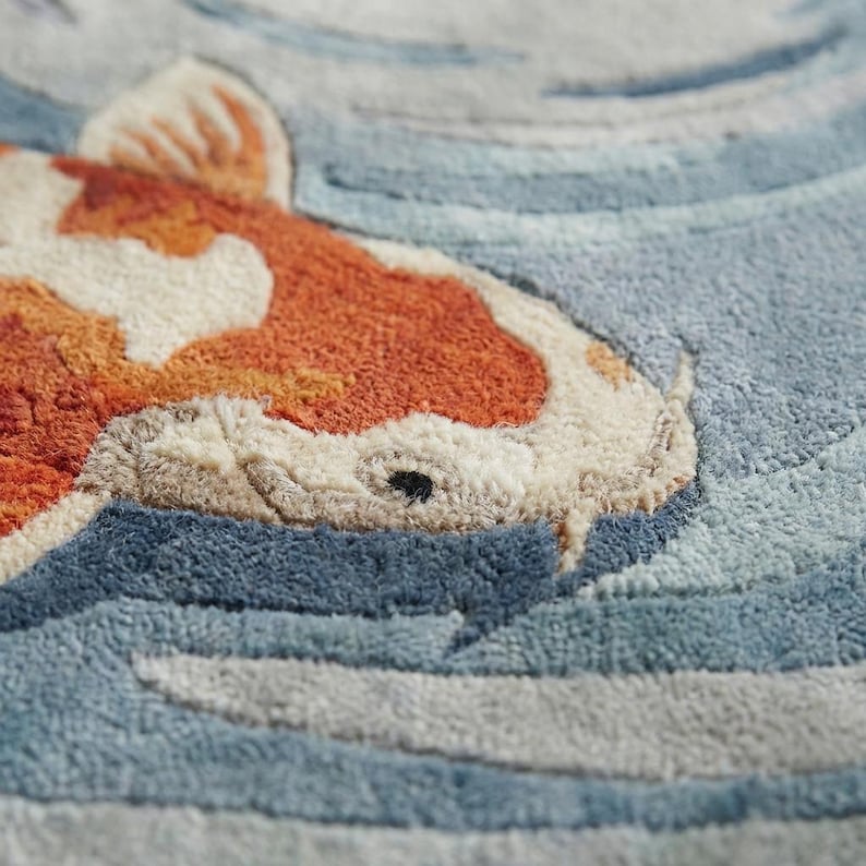 May include: Close-up of a textured rug featuring a koi fish design. The fish has orange and white markings, set against a backdrop of blue and gray water patterns. The rug's texture is visible.
