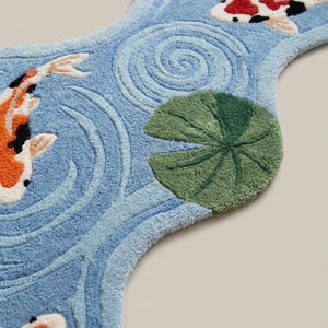Puede incluir: Alfombra azul con forma de arroyo, con peces koi y nen&uacute;fares. Los peces koi tienen marcas naranjas, negras y blancas. La alfombra tiene un aspecto texturizado, con patrones de agua arremolinada y nen&uacute;fares verdes.