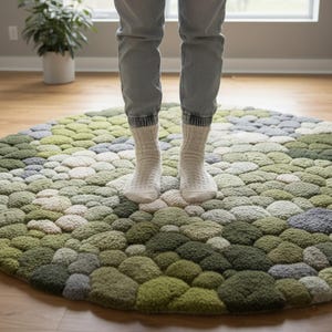 May include: A round rug with a textured, pebble-like design in shades of green, gray, and cream. The rug is on a wooden floor, and a person is standing on it, wearing gray jeans and white socks. The rug's design resembles a natural landscape.