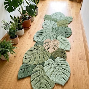 Könnte beinhalten: Ein langer Läufer-Teppich in Form eines Bündels überlappender Monstera-Blätter in verschiedenen Grüntönen, von hellem Salbei bis zu tiefem Waldgrün, mit einem zentralen Blatt in blassem Cremeweiß und detaillierten Blattader-Strukturen.