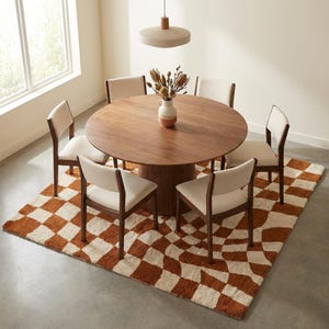 Puede incluir: Una mesa de comedor redonda de madera con seis sillas, colocada sobre una alfombra a cuadros. Las sillas tienen asientos color crema y marcos de madera oscura. Un jarr&oacute;n con flores secas est&aacute; sobre la mesa.