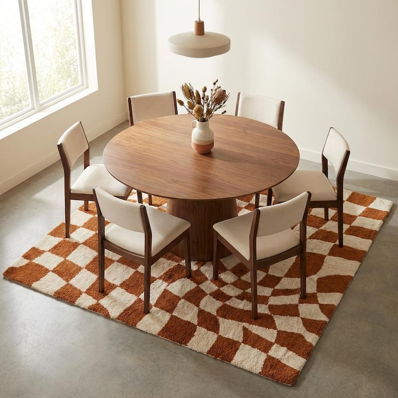 Puede incluir: Una mesa de comedor redonda de madera con seis sillas, colocada sobre una alfombra a cuadros. Las sillas tienen asientos color crema y marcos de madera oscura. Un jarr&oacute;n con flores secas est&aacute; sobre la mesa.
