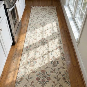 May include: Cream-coloured runner rug with a floral pattern in shades of red, blue, and green. The rug has a fringed edge and is placed on a hardwood floor. The rug's design features stylized flowers and vines.
