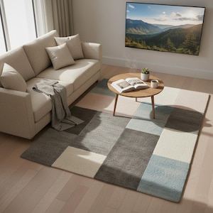 May include: A modern living room featuring a beige sofa with throw pillows, a geometric patterned rug, and a round wooden coffee table. The rug has a patchwork design in shades of gray, blue, and cream. A large landscape painting hangs on the wall.
